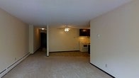 This single-floor residence offers a straightforward and functional layout designed for comfortable everyday living. The main living area is generously open, featuring wall-to-wall carpeting that adds warmth underfoot and a ceiling fan that provides both light and air circulation. The space flows directly into a compact kitchen area, which is equipped with a white electric range, an overhead ventilation hood, and dark cabinetry above — providing ample storage without compromising the open feel of the room. The neutral, cream-toned walls throughout create a clean and versatile backdrop, allowing for a variety of interior design choices. A hallway visible in the background leads to additional areas of the unit, suggesting a practical separation between the living spaces and more private rooms. The overall ambiance is modest and functional, well-suited for those seeking a no-frills, move-in-ready space. The open sightlines between the living and kitchen zones make the interior feel connected and easy to navigate, while the consistent flooring and color palette tie the space together cohesively.