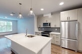 This modern one-bedroom, one-bathroom residence in Framingham, MA offers a well-designed open-concept layout that maximizes functionality and flow. The combined kitchen, dining, and living area serves as the heart of the home, featuring warm wood-tone flooring, crisp white walls, and abundant natural light from large windows with outdoor views of surrounding greenery. The contemporary kitchen is a standout, equipped with stainless steel appliances including a gas range, microwave, and full-size refrigerator, complemented by light wood cabinetry with dark hardware, a gray tile backsplash, and a sleek white quartz island with an undermount sink and stylish pendant lighting overhead. The spacious bedroom is finished with plush carpet and includes access to a walk-in closet outfitted with wire shelving and hanging rods for ample storage. A second closet provides additional organization space. The full bathroom features marble-look tile flooring, a light wood vanity with white countertop, a large mirror, and a storage tower. Additional conveniences include a dedicated laundry area and a utility room. Recessed lighting throughout completes the clean, modern aesthetic of this thoughtfully designed residence.