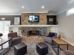 This community room is a thoughtfully designed, single-floor shared space offering a variety of areas to meet residents' social, recreational, and wellness needs. The main lounge area serves as the heart of the space, featuring warm hardwood-style flooring, crown molding, and a striking floor-to-ceiling stone fireplace flanked by built-in shelving. Comfortable seating arrangements, pendant lighting, and curated wall art create an inviting atmosphere for relaxing or socializing. French doors provide access to the outdoors, flooding the space with natural light. Adjacent to the lounge is a dining area furnished with contemporary high-top tables and chairs, illuminated by drum pendant lights and brightened by multiple windows with views of the surrounding grounds. A fully equipped kitchen with rich wood cabinetry, a gas range, microwave, and ample counter space supports catering and resident events. The fitness center is divided into two connected areas, offering cardio equipment including Precor ellipticals and treadmills, as well as strength training machines and a weight bench. Skylights and wall-mounted televisions enhance the workout experience. Throughout the space, neutral tones, crown molding, and consistent hardwood-style flooring tie each area together into a cohesive, polished environment suited for community living.