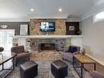 This community room is a thoughtfully designed, single-floor shared space offering a variety of areas to meet residents' social, recreational, and wellness needs. The main lounge area serves as the heart of the space, featuring warm hardwood-style flooring, crown molding, and a striking floor-to-ceiling stone fireplace flanked by built-in shelving. Comfortable seating arrangements, pendant lighting, and curated wall art create an inviting atmosphere for relaxing or socializing. French doors provide access to the outdoors, flooding the space with natural light. Adjacent to the lounge is a dining area furnished with contemporary high-top tables and chairs, illuminated by drum pendant lights and brightened by multiple windows with views of the surrounding grounds. A fully equipped kitchen with rich wood cabinetry, a gas range, microwave, and ample counter space supports catering and resident events. The fitness center is divided into two connected areas, offering cardio equipment including Precor ellipticals and treadmills, as well as strength training machines and a weight bench. Skylights and wall-mounted televisions enhance the workout experience. Throughout the space, neutral tones, crown molding, and consistent hardwood-style flooring tie each area together into a cohesive, polished environment suited for community living.