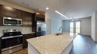 This single-floor unit showcases a thoughtfully designed open-concept layout that seamlessly connects the kitchen and living areas. The kitchen serves as a central highlight, featuring a large granite-topped island with an undermount sink, dark espresso cabinetry extending to the ceiling, and a built-in wine rack. Stainless steel appliances — including a French door refrigerator with ice maker, electric range, and over-the-range microwave — complement the tiled backsplash and matching granite countertops along the perimeter.

Rich hardwood-style flooring flows throughout the main living areas, anchored by recessed lighting and a ceiling fan in the living space. A sliding glass door leads to a balcony, admitting natural light and offering views of a lush, tree-lined landscape visible from multiple windows throughout the unit.

The bedroom features plush carpeting, a ceiling fan, and a large window framing expansive treetop and sky views. The bathroom is finished with large-format tile flooring, a granite-topped dark vanity, and direct access to a walk-in closet with built-in shelving. The overall aesthetic is clean, modern, and cohesive throughout.