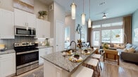 This single-story residence is designed with an open-concept layout that seamlessly connects the kitchen, dining, and living areas, creating a spacious and airy atmosphere enhanced by notably tall ceilings throughout. The kitchen serves as a central feature, outfitted with white shaker cabinetry extending to the upper reaches of the walls, stainless steel appliances including a French door refrigerator, electric range, and built-in microwave. Granite countertops extend across both the perimeter surfaces and a central island, which includes an undermount sink and bar-height seating. A gray tile backsplash adds visual texture, while light wood-style flooring runs throughout the main living areas. Pendant lighting above the island complements the overall design. The living room benefits from generous natural light through large windows and a glass door leading to an outdoor balcony, offering views of the surrounding landscape. A ceiling fan adds to the room's comfort. The bathroom is finished with granite countertops, a single vanity with a framed mirror, and a full soaking tub with tile surround. Adjacent to the bedroom, a walk-in closet provides ample storage with built-in shelving and hanging space.