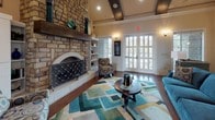 This single-floor space makes a strong architectural impression through its thoughtful blend of rustic and contemporary design elements. The centerpiece of the main lounge area is a soaring floor-to-ceiling natural stone fireplace, complete with a substantial wood mantel and decorative wrought-iron screen, creating a dramatic focal point that anchors the entire room. Exposed dark wood ceiling beams add warmth and character overhead, while rich, polished hardwood floors run throughout, reflecting the ambient light and enhancing the sense of openness.

The lounge is generously proportioned, comfortably accommodating a plush blue velvet sofa, patterned accent chairs, and a round dark wood coffee table set atop a vibrant geometric area rug in shades of teal, green, and gold. Built-in white shelving flanks the fireplace, offering display space and adding a clean, structured contrast to the stone. Natural light pours in through large French doors, which open to views of the surrounding exterior and further brighten the welcoming interior.

Adjacent to the lounge, a compact kitchen or refreshment area continues the stone aesthetic with exposed natural stone walls, warm wood cabinetry, dark countertops, and stainless steel appliances. Wall sconces throughout the space provide soft, layered lighting that complements the overall warm and inviting ambiance.