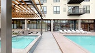This contemporary amenity space is defined by a striking outdoor pool area at its heart, thoughtfully integrated into a multi-story residential building. The pool deck features a resort-style layout with a separate spa, an array of modern chaise lounge chairs, and a wood-beamed pergola that provides both shade and architectural character. The pergola's warm timber elements contrast elegantly with the building's clean, neutral stucco façade and dark steel-framed windows and balconies above.

Floor-to-ceiling glass walls and black-framed doors line the poolside interior, creating a seamless visual connection between the outdoor deck and the indoor common areas. The interiors are finished with large-format light-toned porcelain tile flooring that extends through the corridors, contributing to a clean, polished aesthetic. Recessed lighting illuminates the hallways, which are further enhanced by curated wall art, warm wood-toned doors, and crisp white walls.

Access points feature full-height glass door assemblies with transom windows, flooding entryways with natural light and maintaining sightlines to the pool courtyard. The overall design language is modern and refined, combining durable materials with a cohesive neutral palette that creates a welcoming, upscale ambiance throughout the communal areas.