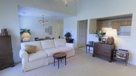 This single-floor space is defined by its dramatic vaulted ceilings, which lend an expansive, airy quality to the main living area. The living room is finished with neutral wall tones and plush carpeting, creating a warm and inviting atmosphere. A classic chandelier serves as a focal point overhead, complementing the traditional aesthetic carried throughout the space. The room comfortably accommodates a large, cream-colored upholstered sofa paired with a decorative round coffee table, while a dark wood dresser along one wall adds both function and character. Framed botanical artwork adorns the walls, adding subtle visual interest. An open pass-through connects the living area to the kitchen, which features warm wood cabinetry and offers a glimpse into an efficiently designed cooking space. A doorway leads to a dining area visible in the background, furnished with seating for several guests. Lush indoor plants are thoughtfully placed throughout, introducing natural color and life. The overall ambiance is clean, traditional, and well-proportioned, with an open flow between living, dining, and kitchen spaces.