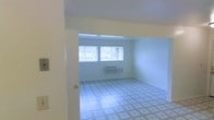 This single-floor space presents a straightforward, functional layout characterized by clean white walls and a patterned tile floor throughout. The main room is an open, rectangular area that benefits from natural light streaming in through two side-by-side windows positioned along the far wall, offering views of the surrounding outdoor environment. The windows contribute to a bright, airy atmosphere despite the modest scale of the space. A wall-mounted HVAC unit is installed beneath the windows, providing a self-contained climate control solution. The ceiling is smooth and white, maintaining the room's clean, utilitarian aesthetic. A standard interior door is visible along one wall, indicating access to an adjacent area or entry point. The patterned tile flooring adds a subtle visual element to the otherwise neutral interior. Overall, the space is unfurnished and move-in ready, presenting a blank canvas that is well-suited for a variety of uses. The simple, no-frills design prioritizes practicality, making it an adaptable and easily maintained environment.
