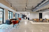Located at 4647 Dodge St. in Omaha, Nebraska, this single-floor coworking space offers a thoughtfully designed environment that blends industrial aesthetics with modern comfort. The expansive open-plan area features polished concrete floors, exposed concrete ceilings with visible ductwork, and track lighting that together create a contemporary industrial ambiance. Large black-framed windows flood the space with natural light and provide views of the surrounding exterior.

The layout accommodates a variety of work styles. Seven private offices, enclosed with glass-panel walls and black metal framing, offer focused workspaces each equipped with a desk and ergonomic seating. Complementing these are multiple lounge areas furnished with bold, colorful seating arrangements — including velvet sofas, leather armchairs, and patterned area rugs — that encourage informal collaboration and relaxation.

A fully equipped kitchen features a sculptural fluted island with a dark countertop, bar seating, open wood shelving, and full appliances with a tiled backsplash. A separate lounge area includes a billiards table and plush seating. Additional communal zones with floor-to-ceiling glass doors connect to outdoor courtyard views. Blue acoustic panels in one corridor add both visual interest and sound management, rounding out a well-appointed, versatile professional environment.