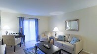 This single-floor residence features a well-appointed living room that serves as its central gathering space. The room is finished with neutral-toned walls and carpeted flooring, creating a warm and inviting atmosphere. A sliding glass door fitted with sheer white vertical blinds and vibrant blue grommet-top curtains allows abundant natural light to fill the space while offering direct access to an outdoor area. The living room is furnished with a plush, light-toned sofa accented by blue throw pillows, complemented by a matching armchair, establishing a cohesive and comfortable seating arrangement. A dark wood coffee table anchors the seating area, while a coordinating side table supports a classic table lamp that adds warm ambient lighting. A framed mirror with a silver finish adorns one wall, enhancing the sense of depth and reflecting light throughout the room. A decorative area rug with blue tones adds visual interest to the floor. The overall ambiance is calm and tastefully styled, balancing functional furnishings with decorative touches that create a welcoming, livable environment.