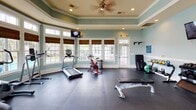 Located at 5806 Ogeechee Rd in Savannah, Georgia, this fitness center is a thoughtfully designed, single-floor facility that combines functionality with an inviting aesthetic. The room features a distinctive tray ceiling with crown molding and soft, sage-green walls accented by crisp white trim, creating a clean and energizing atmosphere. A ceiling fan with recessed lighting ensures comfortable airflow and illumination throughout the space.

One of the most notable architectural features is the expansive arrangement of large, white-framed windows that wrap around much of the room. Dressed with warm bamboo Roman shades, these windows flood the interior with abundant natural light and offer pleasant outdoor views, including glimpses of a pool area and surrounding greenery — a refreshing backdrop for any workout.

The durable dark rubber flooring provides a practical, easy-to-maintain surface suited for a wide variety of exercises. The facility is comprehensively equipped with cardio machines including treadmills, ellipticals, a rowing machine, and stationary and spin bikes. Strength training needs are met with a multi-station cable machine, weight bench, dumbbell rack, medicine balls, and additional functional training accessories. Wall-mounted televisions provide entertainment, enhancing the overall workout experience in this well-appointed fitness space.