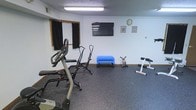 This single-floor fitness room is designed to provide a functional and practical workout environment. The space features durable rubber flooring, which offers both safety and comfort during exercise, and is illuminated by overhead fluorescent lighting that ensures a bright, well-lit atmosphere.

The room is outfitted with a variety of exercise equipment, including a treadmill, a ski-style glider machine, and a dedicated ab workout bench, catering to both cardio and strength training needs. A blue step platform adds versatility for aerobic or low-impact exercises. A wall-mounted flat-screen television serves as an entertainment option to keep users engaged during workouts, while a wall clock helps with timing sessions and intervals.

The walls are painted in a clean, neutral white, giving the space an open and airy feel. A curtained window provides privacy, and framed pictures on the wall contribute a touch of personality to the otherwise utilitarian setting. Wooden baseboards frame the perimeter of the room, adding a subtle warm contrast to the neutral palette. Overall, this fitness room is a straightforward and efficient space thoughtfully arranged to support a range of exercise routines.