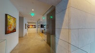 This expansive residential lobby offers a refined first impression through a thoughtfully designed, modern interior. Upon entry, large-format stone and light wood tile flooring transitions seamlessly across zones, anchoring the open layout. A dramatic open-riser staircase with warm wood treads, steel framing, and glass railings serves as a striking architectural focal point, rising beneath soaring double-height ceilings framed by floor-to-ceiling glazing that floods the space with natural light and frames views of a lush landscaped garden courtyard beyond. A sleek reception desk, warmly lit from below and backed by a textured vertical wood slat wall with integrated accent lighting, provides an elegant welcome point. The lounge area features a linear fireplace set into a stone surround, accompanied by contemporary seating arrangements that invite residents to gather in comfort. Curated greenery—including large potted tropical plants—adds life and warmth throughout. A dedicated corridor leads to a bank of elevators and a built-in mailroom, while decorative convex mirrors and carefully selected artwork adorn the walls, contributing to the gallery-like atmosphere. The overall ambiance is one of calm sophistication, blending natural materials, architectural detail, and abundant light into a cohesive, welcoming environment.