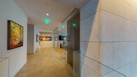 This expansive residential lobby offers a refined first impression through a thoughtfully designed, modern interior. Upon entry, large-format stone and light wood tile flooring transitions seamlessly across zones, anchoring the open layout. A dramatic open-riser staircase with warm wood treads, steel framing, and glass railings serves as a striking architectural focal point, rising beneath soaring double-height ceilings framed by floor-to-ceiling glazing that floods the space with natural light and frames views of a lush landscaped garden courtyard beyond. A sleek reception desk, warmly lit from below and backed by a textured vertical wood slat wall with integrated accent lighting, provides an elegant welcome point. The lounge area features a linear fireplace set into a stone surround, accompanied by contemporary seating arrangements that invite residents to gather in comfort. Curated greenery—including large potted tropical plants—adds life and warmth throughout. A dedicated corridor leads to a bank of elevators and a built-in mailroom, while decorative convex mirrors and carefully selected artwork adorn the walls, contributing to the gallery-like atmosphere. The overall ambiance is one of calm sophistication, blending natural materials, architectural detail, and abundant light into a cohesive, welcoming environment.