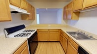 This single-floor space centers around a well-organized kitchen designed for practical, everyday use. The kitchen follows a U-shaped layout, maximizing counter space and workflow efficiency. Warm honey-toned wood cabinetry lines the walls both above and below the counters, providing generous storage throughout. The light-speckled countertops complement the cabinetry and offer ample workspace for meal preparation. A gas range with a black oven is positioned along one wall, accompanied by an overhead range hood, while a stainless steel sink is situated on the opposite side. The light-toned hardwood flooring adds warmth and continuity to the space. A standout architectural feature is the pass-through window centered on the far wall, which opens into an adjacent room, enhancing the sense of openness and allowing natural interaction between spaces. The walls are finished in a clean, neutral tone, keeping the overall atmosphere bright and uncluttered. Recessed lighting provides focused illumination. The kitchen's efficient design and cohesive material palette make it a functional and inviting area within this single-floor space.