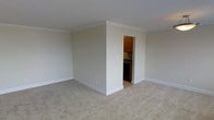 This single-floor space presents a clean, understated interior designed for functional everyday living. The main living area is finished with wall-to-wall beige carpeting that adds warmth underfoot, while neutral-toned walls create a bright, open atmosphere throughout. Crown molding runs along the ceiling perimeter, lending a subtle architectural detail that elevates the overall finish of the room. The walls are painted in a soft, warm beige that complements the light carpet and white trim work, including the clean baseboards lining the floor. Visible through a doorway, the kitchen features warm wood cabinetry in a classic upper-cabinet configuration paired with dark appliances and a simple countertop setup. The transition between the carpeted living space and the kitchen is clearly defined, offering a practical layout. The space benefits from a straightforward, no-frills design aesthetic that prioritizes usability and ease of maintenance. The overall ambiance is calm and neutral, making it a versatile canvas well-suited to a variety of furnishing styles and personal décor preferences.