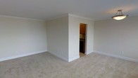 This single-floor space presents a clean, understated interior designed for functional everyday living. The main living area is finished with wall-to-wall beige carpeting that adds warmth underfoot, while neutral-toned walls create a bright, open atmosphere throughout. Crown molding runs along the ceiling perimeter, lending a subtle architectural detail that elevates the overall finish of the room. The walls are painted in a soft, warm beige that complements the light carpet and white trim work, including the clean baseboards lining the floor. Visible through a doorway, the kitchen features warm wood cabinetry in a classic upper-cabinet configuration paired with dark appliances and a simple countertop setup. The transition between the carpeted living space and the kitchen is clearly defined, offering a practical layout. The space benefits from a straightforward, no-frills design aesthetic that prioritizes usability and ease of maintenance. The overall ambiance is calm and neutral, making it a versatile canvas well-suited to a variety of furnishing styles and personal décor preferences.