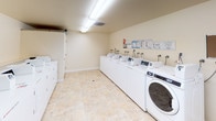 This shared laundry facility is a practical, single-floor space designed to accommodate the needs of multiple users efficiently. The room features a generous row of commercial-grade front-loading and top-loading washers and dryers from the Speed Queen and WASH WAM lines, all neatly arranged along the walls to maximize usable floor space. Instructional and informational notices are posted above the machines for easy reference. The flooring throughout is a durable, stone-patterned tile in warm neutral tones, contributing to the clean and welcoming atmosphere of the room. Overhead fluorescent lighting ensures the space is well-lit at all hours. A centrally positioned folding table provides a convenient surface for sorting and folding laundry. A window with a multi-pane black frame allows natural light to enter and offers a view of the surrounding greenery outside. Additional practical features include a cork bulletin board for community notices, a fire extinguisher mounted near the exit, and multiple utility connections along the walls for the machines. The space is finished with smooth cream-colored walls and white baseboards, giving the room a tidy, maintained appearance. The layout is open and spacious, allowing comfortable movement between the machines and the folding area.