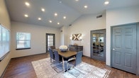 This conference room offers a polished and functional environment designed to accommodate productive meetings and collaborative sessions. The space immediately impresses with its dramatic vaulted ceiling, which lends an open, airy quality rarely found in professional meeting spaces. Recessed lighting is thoughtfully distributed throughout, providing consistent, well-balanced illumination across the entire room.

The centerpiece of the space is a substantial dark wood conference table surrounded by six contemporary gray upholstered chairs with sleek chrome legs, all positioned atop a neutral, textured area rug over rich hardwood flooring. Multiple windows fitted with horizontal blinds line two walls, allowing natural light to fill the room while offering adjustable privacy and glare control. The views to the exterior greenery contribute a calm, pleasant atmosphere.

Neutral gray walls provide a clean, professional backdrop, complemented by dark-trimmed doors and window frames that add visual definition. A glass-paned interior door offers a sightline into an adjacent workspace, while a separate exterior door provides convenient access. A sideboard with decorative lighting anchors one wall, adding a refined finishing touch. The overall ambiance is modern, welcoming, and well-suited for focused professional gatherings.