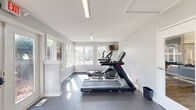 Located at 4365 Bayshire Rd. in Groveport, OH, this single-floor fitness center offers a well-appointed and efficiently designed workout space. The room is finished with clean white walls and light gray tones, creating a bright, modern atmosphere that feels open and inviting. Durable rubber flooring throughout provides a practical, slip-resistant surface suited for a range of exercise activities.

Large multi-pane windows line the walls on multiple sides, flooding the space with abundant natural light and offering pleasant views of the surrounding outdoor greenery. A set of glass-paneled double doors serves as the main entry and exit point, marked by an illuminated exit sign above, and a keypad access system ensures secure entry.

The fitness center is equipped with a thoughtful selection of cardio machines, including treadmills, an elliptical, and a stationary cycle — all from Star Trac. A wall-mounted flat-screen television provides entertainment during workouts. On the strength-training side, a tiered dumbbell rack with a range of free weights and a weight bench offer options for resistance training. A large wall mirror on one end amplifies the sense of space and allows users to monitor their form. Overhead linear fluorescent lighting supplements natural light, ensuring the space remains well-lit at all times. An interior glass-panel door connects to an adjacent area, adding to the overall functionality of this compact but complete fitness facility.