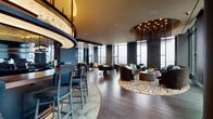 This upscale rooftop lounge is a masterclass in contemporary interior design, combining dramatic architectural elements with a refined, moody aesthetic. Soaring floor-to-ceiling windows wrap the space, flooding it with natural light while framing expansive panoramic city views that serve as a stunning living backdrop throughout the day.

The open-plan layout is anchored by a striking curved bar with a sleek countertop, complemented by upholstered bar seating in deep navy tones. Overhead, an eye-catching circular chandelier with cascading fringe-like elements adds warmth and drama to the lounge area below, where a generously sized curved sofa, plush armchairs, and a richly patterned area rug create an inviting social hub.

Rich dark hardwood flooring runs throughout, grounding the space and contrasting beautifully with the dark walls and warm gold ceiling accents. A dedicated seating nook features bold geometric décor pieces in gold tones alongside velvet accent chairs, while a game or meeting table with leather seating offers a more structured gathering option. A decorative tile feature wall with intricate patterning adds textural interest to one corner.

Direct access to a spacious outdoor terrace expands the experience further, providing an alfresco setting with additional seating. The overall ambiance is sophisticated, intimate, and elevated—ideal for upscale social gatherings.
