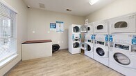 Located at 100 Alexander Xing in Loganville, GA, this self-contained laundry facility offers a practical and well-organized space designed for efficient use. The room features light-toned walls and durable wood-look flooring that contribute to a clean, low-maintenance environment. Multiple stacked washer and dryer units line the walls, providing ample laundry capacity within a compact footprint. The machines are equipped with digital controls and app-compatible payment systems, reflecting a modern approach to shared laundry amenities. A built-in folding counter with a laminate top offers a convenient surface for sorting and folding laundry. Natural light enters through windows fitted with horizontal blinds, brightening the interior and providing views of the surrounding outdoor greenery. Overhead ventilation registers ensure proper airflow throughout the space. An electrical panel and a wall-mounted fire extinguisher are clearly visible, underscoring attention to safety. Informational signage is posted throughout the room, offering guidance on machine use and app-based laundry tracking. A waste bin is conveniently positioned near the folding area. The single-floor layout is straightforward and accessible, making this facility a functional and user-friendly laundry resource for the community it serves.