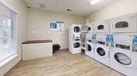 Located at 100 Alexander Xing in Loganville, GA, this self-contained laundry facility offers a practical and well-organized space designed for efficient use. The room features light-toned walls and durable wood-look flooring that contribute to a clean, low-maintenance environment. Multiple stacked washer and dryer units line the walls, providing ample laundry capacity within a compact footprint. The machines are equipped with digital controls and app-compatible payment systems, reflecting a modern approach to shared laundry amenities. A built-in folding counter with a laminate top offers a convenient surface for sorting and folding laundry. Natural light enters through windows fitted with horizontal blinds, brightening the interior and providing views of the surrounding outdoor greenery. Overhead ventilation registers ensure proper airflow throughout the space. An electrical panel and a wall-mounted fire extinguisher are clearly visible, underscoring attention to safety. Informational signage is posted throughout the room, offering guidance on machine use and app-based laundry tracking. A waste bin is conveniently positioned near the folding area. The single-floor layout is straightforward and accessible, making this facility a functional and user-friendly laundry resource for the community it serves.