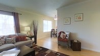 This single-floor residence offers a welcoming and well-appointed living space defined by a cohesive open-concept layout that seamlessly connects the living and dining areas. The interior is finished with warm neutral walls and light hardwood flooring that runs throughout, creating a bright and airy atmosphere.

The living area is thoughtfully arranged with comfortable seating, including a sofa and a wicker accent chair adorned with decorative pillows in warm earth tones. A richly patterned area rug anchors the space, while a side table with a classic lamp adds a cozy, layered ambiance. Decorative elements such as tall branch arrangements and framed botanical artwork contribute to the space's approachable, inviting character.

Crown molding traces the ceiling lines, adding an elegant architectural detail that elevates the overall aesthetic. A ceiling fan provides both function and visual interest above the dining area, which is furnished with a dark wood table and chairs and receives generous natural light through a large window fitted with horizontal blinds.

The overall ambiance is warm, livable, and tastefully decorated, striking a balance between comfort and style. The open flow between living and dining areas makes the space feel expansive and well-suited for both everyday living and casual entertaining.