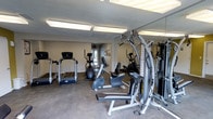 This fitness center offers a practical and functional workout environment designed to accommodate a variety of exercise routines. The space is outfitted with a selection of cardio equipment, including treadmills, an elliptical trainer, and a stationary recumbent bike, providing users with multiple options for cardiovascular training. A multi-function cable strength machine is positioned along one side, enabling a range of resistance-based exercises.

Full-length mirrors line one wall, visually expanding the space and allowing users to monitor their form during workouts. Overhead fluorescent lighting ensures the area is well-lit throughout. The neutral-toned walls and light flooring create a clean, no-frills atmosphere focused on utility. A window near the back of the room allows for a degree of natural light, contributing to a more open feel. The equipment is arranged to allow comfortable movement between stations, making efficient use of the available floor space. Overall, the fitness center provides a straightforward and accessible environment for residents or guests seeking a convenient place to maintain their fitness routines.