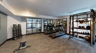 Located at 320 Second Ave S in Kirkland, WA, this well-appointed amenity facility spans a single floor and is divided into three distinct areas designed to serve both residents and their pets. The fitness center forms the heart of the space, offering a generously sized workout area outfitted with treadmills, elliptical machines, stationary bikes, strength training equipment, and a cable machine system. Large black-framed glass partition walls create defined zones within the gym while maintaining an open, airy feel. Neutral-toned carpet flooring, recessed lighting, and wall-mounted mirrors contribute to a clean, modern aesthetic throughout. A bold motivational mural — featuring a neon "Don't Quit" sign set against dynamic fitness imagery — energizes one accent wall, lending the space an encouraging atmosphere. Natural light filters in through multiple windows, adding brightness to the workout areas. A separate pet spa room rounds out the facility, thoughtfully designed with large-format tile flooring, a built-in pet wash basin, a grooming table, and a charming dog-silhouette mural with a green tile accent border. The entry corridor features glass doors with frosted panels and clear "Fitness Center" signage, providing a polished, welcoming introduction to the space.