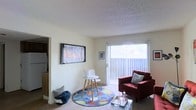 This single-floor residence offers a comfortable and functional layout designed for everyday living. The living room serves as the heart of the space, featuring wall-to-wall carpeting and a textured ceiling that adds subtle character. A large sliding glass door with vertical blinds dominates one wall, flooding the room with natural light and providing access to an outdoor area with open views. The living area is thoughtfully arranged, with ample room for seating and a wall-mounted flat-screen television, making it an inviting space for relaxation and entertainment. A colorful circular area rug anchors the seating arrangement and adds a lively pop of color to the neutral tones of the room. Adjacent to the living space, an open passageway leads directly into the kitchen, which is visible in the background and features standard appliances including a refrigerator. The overall ambiance of the residence is light and airy, with neutral-toned walls and natural light contributing to a welcoming atmosphere throughout. The straightforward, open flow between the main living and kitchen areas makes the space feel connected and easy to navigate.