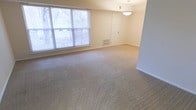 This single-floor space offers a clean, straightforward layout designed for comfortable everyday living. The main living area is generously proportioned, featuring wall-to-wall neutral beige carpeting that lends warmth and a soft, inviting feel underfoot. A large multi-pane window dressed with horizontal blinds dominates one wall, allowing an abundance of natural light to fill the room while offering views of the surrounding greenery outside. The interior walls are finished in a soft, creamy white tone that complements the light palette throughout. A classic pendant light fixture hangs from the ceiling, providing a central focal point and functional overhead illumination. A floor-level HVAC vent is integrated into the wall, ensuring efficient climate control. The room flows naturally toward a doorway leading to additional areas of the space. The overall ambiance is calm, neutral, and move-in ready, making it a versatile canvas well-suited for a variety of furniture arrangements and interior styles. The combination of ample natural light, open floor space, and understated finishes creates an atmosphere that feels both practical and welcoming.