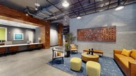 This thoughtfully designed single-floor commercial space combines industrial and contemporary aesthetics to create a dynamic, multi-functional environment. Polished concrete floors extend throughout the open layout, anchoring the space with a clean, urban foundation. Reclaimed wood accents on columns, ceiling panels, and wall cladding introduce warmth that balances the cool gray brick walls and exposed ductwork overhead.

The reception area features a long, low-profile counter with seating for guests, framed by colorful wall art and warm recessed lighting beneath a wood-paneled ceiling. Adjacent to it is a refreshment station with built-in cabinetry, providing a welcoming point of arrival.

The lounge area is a standout zone, centered on a vibrant blue area rug layered with a cognac leather sofa, mid-century-style armchairs, round wood coffee tables, and chartreuse ottomans. A large mixed-media artwork commands the gray brick feature wall, complemented by pendant lighting suspended from a black grid ceiling structure.

A glass-walled conference room offers floor-to-ceiling windows with expansive street views, a generous wood-top meeting table, and a wall-mounted display screen — ideal for collaborative sessions. A private workstation tucked along a corridor provides a quiet, focused work setting with a built-in desk and additional wood accent detailing. The space balances openness with intimate functionality throughout.