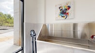 This dedicated pet grooming space is thoughtfully designed with both functionality and aesthetics in mind. The room centers around a large, professional-grade stainless steel grooming tub equipped with built-in plumbing fixtures, a retractable ramp for easy pet access, and a stainless steel backsplash that extends to a convenient shelf above. The walls are finished with a clean, modern hexagonal tile treatment on the lower half, transitioning to crisp white painted surfaces above, creating a bright and hygienic environment. White cabinetry positioned above the tub provides additional storage. The light-toned hexagonal floor tiles complement the overall clean aesthetic and are well-suited for a wet environment. A frosted glass interior door offers privacy while maintaining a sense of openness, and a full-length glass exterior door floods the room with natural light and provides direct access to an outdoor courtyard with landscaping and iron fencing. A professional-grade pet blow dryer on a mobile stand rounds out the grooming equipment. A vibrant, colorful canvas print of a dog adds a cheerful, welcoming touch to the space, reinforcing its pet-friendly purpose.