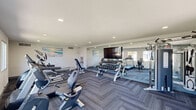 Located at 3330 West Greenway Road in Phoenix, Arizona, this fitness center offers a well-appointed exercise room designed to support a comprehensive workout routine. The space features a herringbone-patterned carpet floor, recessed lighting, and a textured white ceiling that contribute to a clean, modern aesthetic. Large windows along multiple walls allow natural light to fill the room and provide views of the surrounding outdoor area.

The exercise room is equipped with a wide variety of cardio and strength-training equipment. Cardio offerings include treadmills, an elliptical, a stationary bike, and a rowing machine, all from the Spirit brand. A full cable and pulley multi-functional strength machine is positioned near a floor-to-ceiling mirror wall, which visually expands the space and supports form monitoring during workouts. A dumbbell rack with a full range of free weights, a flat bench, and an adjustable weight bench round out the strength training options. Exercise balls in multiple sizes are also available for flexibility and core training.

A wall-mounted flat-screen television provides entertainment during workouts, while a water fountain offers convenient hydration. Coastal-themed artwork adds a relaxed, inviting touch to the otherwise functional environment, making this fitness center a practical and welcoming space for daily exercise.