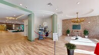 Located at 30 W Broad St in downtown Bethlehem, PA, this generously sized single-floor amenity space offers a thoughtfully designed environment that blends social, recreational, and lounge functions. The entrance lobby greets visitors with light hardwood flooring, exposed brick accent walls, a vintage film projector display, and floor-to-ceiling windows that flood the space with natural light while framing views of the surrounding streetscape. Architectural details such as arched niches with built-in shelving, herringbone-patterned hardwood floors, and a curved ceiling with cove lighting add visual interest throughout. A spacious game room features a pool table, shuffleboard table, and a striking double-sided tiled fireplace, all anchored by modern pendant lighting. Multiple lounge areas are furnished with sectional sofas, patterned area rugs, wall-mounted televisions, and curated décor including colorful abstract artwork displayed against sage green accent walls. A dedicated dining and social area with a wet bar and custom shelving provides an ideal space for gatherings. Large sliding glass doors in one lounge area open to an outdoor terrace, seamlessly extending the living space. The overall ambiance is modern yet welcoming, combining contemporary finishes with warm, eclectic touches throughout.