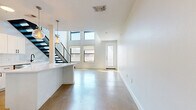 This contemporary two-story residence is thoughtfully designed across two equally sized floors, offering a modern aesthetic highlighted by clean lines and quality finishes throughout. The main level makes an immediate impression with its soaring double-height ceilings and an abundance of natural light streaming through a dramatic arrangement of stacked windows flanking the entry door. Polished concrete floors run throughout the ground floor, lending an industrial-chic character to the open living and kitchen areas. A bold open-riser staircase with sleek black metal railings and cable detailing serves as a striking architectural focal point. The kitchen is well-appointed with white shaker cabinetry, subway tile backsplash, stainless steel appliances, a white quartz island with storage, and globe pendant lights that add a warm, contemporary glow. The bathroom on the lower level features full white subway tile walls with a soaking tub and separate shower, accented by matte black fixtures. Upstairs, the second floor transitions to warm carpeting and features an open loft-style layout with a ceiling fan, a secondary bathroom with wood-look flooring, white cabinetry, and matte black hardware. The upper level overlooks the main living area below through the black metal railing, reinforcing the home's cohesive, modern design language.