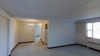 This single-floor residence presents a clean, well-maintained interior with a straightforward and functional layout. The main living area is open and airy, featuring light-colored walls and dark baseboards that frame the space with subtle contrast. Crown molding runs along the upper edges of the walls, adding a classic architectural touch to the room. The flooring transitions between carpet in the living area and what appears to be a hard surface in the adjoining spaces, creating natural zone distinctions throughout the home. A standout feature is the elegant gold-toned chandelier that illuminates the transitional area connecting the living space to the rest of the home, casting a warm and inviting glow. This hallway or pass-through area leads further into the interior, hinting at additional rooms beyond. A white refrigerator is visible near the transition into the kitchen area, suggesting a practical galley-style or compact kitchen nearby. The overall ambiance is calm and neutral, with a palette of soft whites and warm beige tones that lend the space a bright, welcoming feel. The layout appears to flow efficiently from the living area through to the kitchen and adjoining rooms, making this a comfortable and cohesive single-level home.
