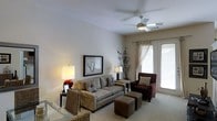 This single-floor space presents a clean, open layout designed for both comfort and functionality. The main living area is generously proportioned, featuring soft, neutral-toned walls that contribute to a bright and airy atmosphere. Plush carpeting covers the floor throughout the primary living space, adding warmth underfoot, while a transition to what appears to be granite or terrazzo flooring is visible at the foreground, suggesting a distinct functional zone such as a kitchen or entryway. A set of elegant French doors at the far end of the room floods the interior with natural light and provides direct access to the outdoors, enhancing the sense of openness. Overhead, a ceiling fan with integrated lighting ensures year-round comfort, while a stylish track lighting system with multiple adjustable fixtures adds focused illumination and a modern touch to the space. The clean white ceilings and minimal architectural detailing give the interior a timeless, move-in-ready quality. The overall ambiance is calm and inviting, making the space well-suited for a variety of uses and interior design styles.