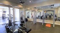 This single-story fitness center offers a comprehensive workout environment designed to accommodate a range of strength training and exercise needs. The space is outfitted with multiple cable machine stations, a barbell rack loaded with weight plates, and adjustable workout benches, providing users with versatile options for both resistance and free weight training. The flooring consists of durable rubber matting, which is well-suited for heavy equipment use and provides safety and stability during workouts. Large mirrors line the walls, visually expanding the space while allowing users to monitor their form during exercise. Ceiling fans are mounted overhead, contributing to air circulation and comfort throughout the room. Recessed lighting in the ceiling, combined with natural light filtering in through windows visible in the background, ensures the space is well-lit and inviting. The neutral, light-toned walls and open layout create an uncluttered, functional atmosphere. Overall, the fitness center is thoughtfully arranged to maximize usable floor space while maintaining an organized and accessible environment for users of varying fitness levels.