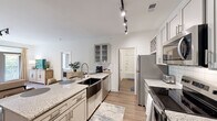 Located at 9051 Seneca Dr in Wilmington, NC, this thoughtfully designed one-bedroom, two-bathroom residence offers a well-appointed single-floor layout with modern finishes throughout. The open-concept kitchen and living area serves as the heart of the home, featuring an expansive granite island with bar seating, a deep farmhouse sink, stainless steel appliances including a refrigerator, range, and built-in microwave, and white subway tile backsplash. Track lighting illuminates the space, and gray-toned cabinetry with glass-front upper cabinets adds a refined touch. The living room offers warm wood-look flooring, ample natural light, and a relaxed coastal ambiance. The bedroom is carpeted for comfort, features a ceiling fan, large windows with garden views, and direct access to a private balcony. A generous walk-in closet with built-in shelving connects to one of the two full bathrooms, which includes a tub-shower combination with white tile surround. The second bathroom is conveniently accessible from the main living area. Clean white walls, consistent finishes, and a cohesive coastal aesthetic make this residence a comfortable and stylish home in the Wilmington area.