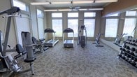 This fitness center in Midland is designed to support an active lifestyle, offering a thoughtfully arranged single-floor workout space equipped with a variety of exercise machines. The room is bathed in natural light, courtesy of multiple large windows along the walls, including a trio of tall, prominent windows at the front that provide views of the surrounding neighborhood. Additional transom windows near the ceiling further enhance the brightness of the space.

The fitness center is outfitted with two treadmills, an elliptical trainer, a stationary bike, and a seated strength-training machine, providing options for both cardiovascular and resistance-based workouts. The equipment is arranged to allow comfortable movement throughout the room.

A ceiling fan is centrally mounted overhead, contributing to air circulation and comfort during workouts. The floor is covered in a patterned, low-pile carpet that adds a subtle decorative element while providing a stable surface underfoot. The neutral-toned walls and clean layout give the space a practical yet welcoming atmosphere, making it a functional and accessible fitness facility.