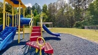 Located at 512 Nottingham Ct in Hopewell, Virginia, this outdoor community amenity area offers a well-rounded set of recreational features designed to serve residents of all ages. At the heart of the space is a colorful, multi-component playground structure equipped with slides, climbing stairs, and interactive play elements, all set atop a gravel safety surface for added protection. A pergola sits nearby on a lush open lawn, providing a shaded gathering spot framed by mature trees that line the perimeter and create a naturally scenic backdrop. Adjacent to the playground, a fenced dog park with a dedicated green space offers a secure area for pets to roam freely. A community swimming pool is visible in the background, adding another layer of recreational value to the grounds. The well-maintained open lawn areas provide generous space for outdoor activities and community gatherings. A sizable paved parking lot with clearly marked spaces ensures convenient access for residents and guests. The surrounding residential buildings, consistent in their neutral exterior finishes, contribute to the cohesive character of the neighborhood. Abundant natural light, open skies, and the presence of tall trees throughout create a welcoming, park-like atmosphere that enhances the overall quality of this community outdoor space.