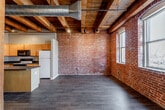 Located on Berkley Pkwy in Kansas City, MO, this single-floor loft showcases a compelling industrial aesthetic rooted in its historic building origins. Throughout the space, exposed red brick walls serve as a defining architectural element, appearing in the main living areas, kitchen, bathroom, and laundry room, lending warmth and texture to every room.

The open-plan living and kitchen area is anchored by heavy timber ceiling beams and structural wooden columns, complemented by visible ductwork that reinforces the loft's industrial character. Dark hardwood flooring runs continuously through these spaces, creating a cohesive and grounded feel. The kitchen features light wood cabinetry, dark granite-style countertops, a central island, and a full suite of appliances, all beneath the striking exposed beam ceiling with track lighting.

The bedroom area offers carpeted flooring for comfort, exposed beam ceilings with a ceiling fan, and painted walls that soften the industrial elements. The bathroom continues the brick wall theme alongside a wood vanity with dark countertop and a tub-shower combination. A dedicated laundry area with a full-size washer and dryer adds practical convenience. Natural light enters through windows, enhancing the overall warmth of this character-rich urban space.