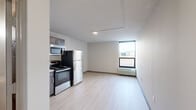 This efficiently designed single-floor studio offers a clean, contemporary layout that maximizes its compact footprint. The open-plan living and kitchen area is anchored by light-toned, wood-style flooring that runs throughout, creating a cohesive and bright atmosphere. A textured ceiling adds subtle dimension overhead.

The kitchen is well-appointed with dark espresso cabinetry, light countertops, and a full suite of stainless steel appliances, including a refrigerator, electric range, over-the-range microwave, and a single-basin sink — offering complete functionality within a streamlined space.

The living area extends toward a large picture window framed in dark trim, which draws in natural light and provides views of the surrounding exterior. A wall-mounted PTAC heating and cooling unit sits beneath the window, ensuring year-round climate control.

The bathroom contrasts with the rest of the unit through its warm, earth-toned tile work, featuring a decorative diamond-pattern border, a granite-topped vanity with an undermount sink, and a tiled shower area with a towel rack — combining practicality with a more traditional aesthetic finish.