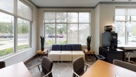 This professionally designed business center offers a well-appointed, single-floor environment tailored to support productivity and collaboration. The space is organized into distinct functional zones that flow seamlessly into one another, creating a cohesive and polished atmosphere throughout.

A dedicated workstation counter runs along a large window wall, fitted with a granite-style surface, under-counter stools, pendant globe lighting, and essential office equipment including a printer and coffee station — providing everything needed for focused individual work while maintaining views of the surrounding greenery outside.

Adjacent to the workstation area, a comfortable lounge zone features modular seating — including cream ottomans, a navy accent sofa, and leather arm chairs — flanked by tall potted snake plants and framed by floor-to-ceiling windows that flood the space with natural light.

A semi-private conference area accommodates small group meetings with wooden tables and sleek grey chairs, set against a striking textured blue accent wall framed in warm wood trim. A large flat-screen display is mounted on the accent wall, ideal for presentations.

Decorative elements include a bold abstract large-format artwork, a sheer blue floor-to-ceiling panel partition, and a patterned carpet in neutral tones that ties all zones together. An elegant chandelier and recessed lighting complete the refined, contemporary ambiance of this fully equipped business center.