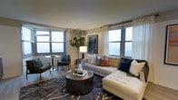 Located at 1303 Delaware Ave in Wilmington, Delaware, this single-floor residence offers a refined and contemporary living environment. The living area serves as the centerpiece of the space, showcasing a well-appointed layout designed for both comfort and functionality.

Large windows on multiple walls flood the room with natural light, framing views of the surrounding Wilmington skyline and creating an airy, open atmosphere. Light-toned walls complement the wood-style flooring, contributing to a clean and modern aesthetic throughout.

The living room is generously sized, accommodating a large tufted sectional sofa accented with an assortment of decorative pillows in neutral, navy, and terracotta tones. A dark, industrial-style round coffee table anchors the seating area atop a patterned area rug, adding texture and visual interest. A compact dining nook is situated near the windows, featuring a round bistro-style table and chairs — ideal for casual meals or working from home.

Decorative touches, including a tall indoor tree and framed wall art, bring warmth and personality to the space. The overall ambiance is sophisticated yet inviting, blending modern design with comfortable, livable style.