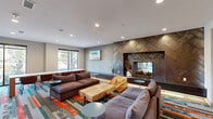 Located at 1033 East Sawmill Road in Flagstaff, Arizona, this thoughtfully designed community living room offers a welcoming and multifunctional gathering space across a single floor. The room's most striking architectural feature is a floor-to-ceiling fireplace wall clad in rich, metallic herringbone-patterned tile, creating a dramatic focal point that anchors the lounge area. Complementing this is an adjacent textured stone accent wall, adding depth and warmth to the overall aesthetic.

The lounge seating area is furnished with plush sofas and accent chairs arranged around a live-edge wood coffee table, encouraging relaxed conversation and comfort. Recessed lighting throughout the ceiling provides consistent, even illumination, while large sliding glass windows and doors invite generous natural light and views of the surrounding Flagstaff landscape.

A vibrant, multi-colored patterned carpet in bold tones of teal, orange, red, and gray runs throughout the space, tying the room's modern design together with an energetic, southwestern-inspired character. A dedicated workspace area along one wall features a built-in desk with computer workstations, cabinetry, and a central collaborative table with seating, making the space equally suited for productivity. Glass-paneled doors provide visual connectivity to adjacent areas, enhancing the open and accessible feel of this well-appointed community space.