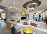 Located at 405 NE 2nd St in Florida, this single-floor amenity space combines contemporary design with practical fitness facilities. The standout feature is a sophisticated lounge area anchored by a striking ceiling installation of interlocking circular LED light fixtures set against a dark recessed panel, creating a bold visual focal point. The lounge is furnished with a curved white sectional sofa accented by colorful throw pillows, a gold accent table, and stylish cage-style chairs. A large-scale city map mural branded "The Rise" serves as a prominent feature wall, lending the space a distinct urban identity. The open layout flows naturally between seating zones, with large windows drawing in natural light and views of the surrounding greenery.

The fitness center is well-appointed with a comprehensive selection of commercial-grade cardiovascular equipment, including multiple treadmills, an elliptical, a stationary bike, and a stair climber. A dedicated strength-training area provides a range of selectorized resistance machines suited for full-body workouts. Large windows and glass doors throughout the fitness space offer outdoor views and abundant natural light, contributing to an energizing workout environment. The durable rubberized flooring and clean, bright interiors maintain a professional and functional atmosphere throughout.
