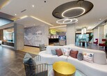 Located at 405 NE 2nd St in Florida, this single-floor amenity space combines contemporary design with practical fitness facilities. The standout feature is a sophisticated lounge area anchored by a striking ceiling installation of interlocking circular LED light fixtures set against a dark recessed panel, creating a bold visual focal point. The lounge is furnished with a curved white sectional sofa accented by colorful throw pillows, a gold accent table, and stylish cage-style chairs. A large-scale city map mural branded "The Rise" serves as a prominent feature wall, lending the space a distinct urban identity. The open layout flows naturally between seating zones, with large windows drawing in natural light and views of the surrounding greenery.

The fitness center is well-appointed with a comprehensive selection of commercial-grade cardiovascular equipment, including multiple treadmills, an elliptical, a stationary bike, and a stair climber. A dedicated strength-training area provides a range of selectorized resistance machines suited for full-body workouts. Large windows and glass doors throughout the fitness space offer outdoor views and abundant natural light, contributing to an energizing workout environment. The durable rubberized flooring and clean, bright interiors maintain a professional and functional atmosphere throughout.
