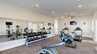 This fitness center is a thoughtfully designed, single-floor facility offering a comprehensive range of workout equipment and dedicated exercise zones. The space is divided into two primary areas, each serving distinct training purposes and connected by an open layout that promotes easy movement throughout.

The main workout area features a generous selection of cardio equipment, including multiple treadmills and elliptical trainers, each positioned for ample personal space during use. A wall-mounted flat-screen television is installed for entertainment during cardio sessions. A ceiling fan adds to the comfort of the room. Large wall mirrors line one side of the space, visually expanding the room and allowing users to monitor their form during exercise.

A dedicated free weights section houses a full rack of dumbbells spanning a wide range of sizes, accompanied by a weight bench, making it ideal for strength training routines. Stationary cycling bikes and additional cardio machines round out the equipment offerings, catering to a variety of fitness preferences.

The flooring consists of durable, textured carpet throughout the main exercise areas, providing cushioning and traction. Recessed lighting and natural light from windows keep the environment bright and welcoming. The neutral-toned walls and clean white trim contribute to a fresh, well-maintained atmosphere. An entryway area with a patterned rug offers a transitional space upon entry, and a separate room provides additional storage or supplementary workout space.