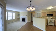 This single-floor space offers an open and functional layout anchored by a welcoming living area. A white tile-surround fireplace serves as the focal point of the room, flanked by built-in wall shelves that provide both display and storage options. Crown molding runs along the ceiling, adding a refined architectural detail throughout. An elegant black wrought-iron chandelier with frosted glass shades hangs centrally, complementing the recessed lighting for layered illumination. The flooring transitions from warm hardwood in the main traffic areas to plush carpet in the living space, clearly defining zones while maintaining a cohesive aesthetic. Natural light filters in through a window fitted with white blinds. The space flows openly toward the kitchen, which is partially visible through a pass-through counter with a raised bar-height ledge — ideal for casual dining or conversation. Light wood cabinetry is visible in the kitchen, contributing to the overall bright and neutral color palette of warm beige tones and white trim. A hallway leading to additional rooms is accessible from the main living area, completing this well-connected, single-level layout.